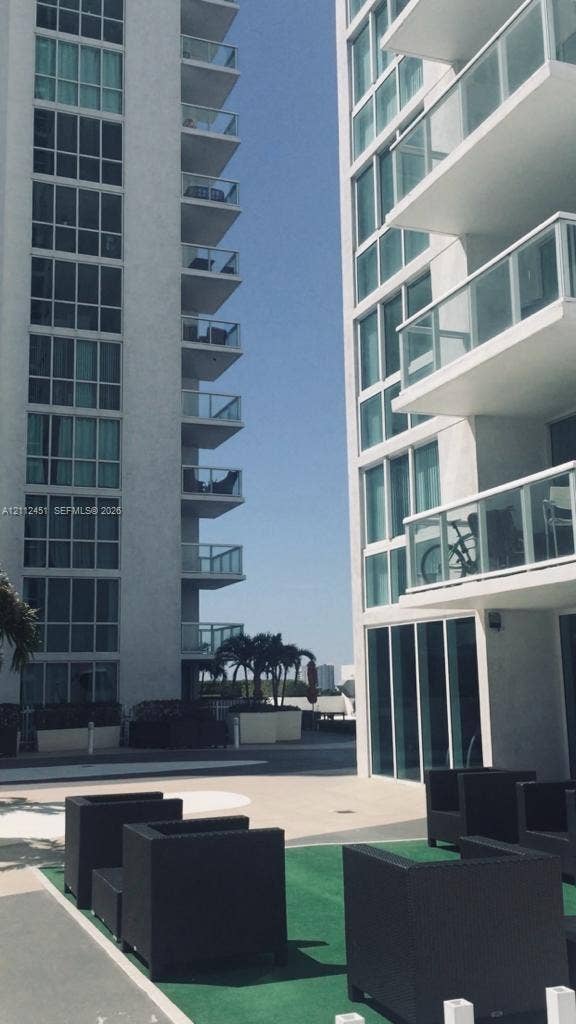 High-rise exterior featuring floor-to-ceiling glass windows, stacked private balconies with glass railings, and a concrete-paved outdoor terrace area