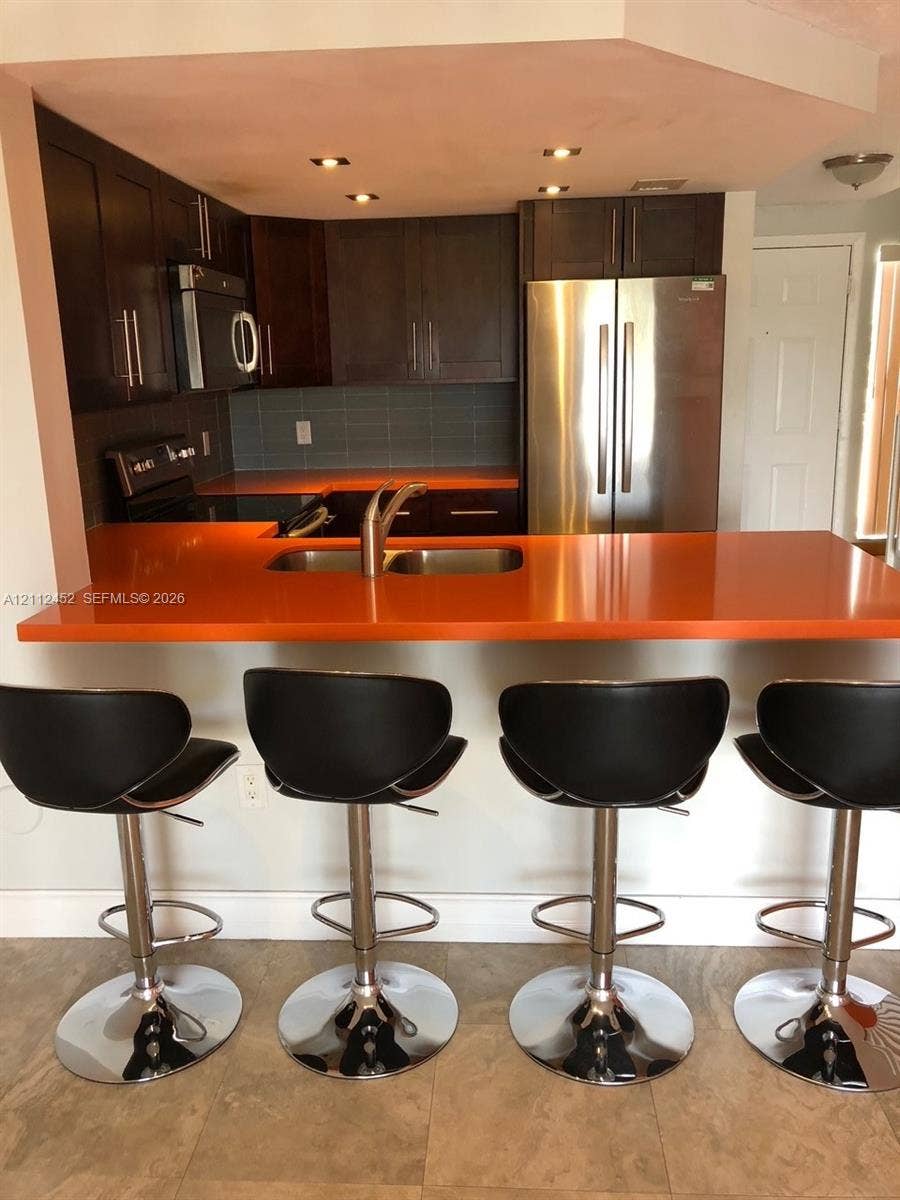 Kitchen featuring dark cabinetry, stainless steel appliances, solid-surface peninsula countertop, undermount sink with arched faucet, and tiled backsplash