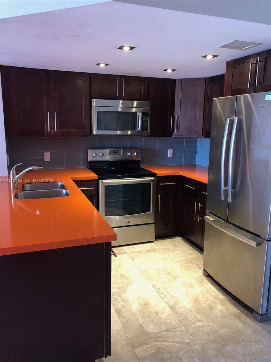 Kitchen featuring dark wood-finish cabinetry, bright solid-surface countertops, stainless steel appliances, recessed ceiling lighting, and large-format tile flooring