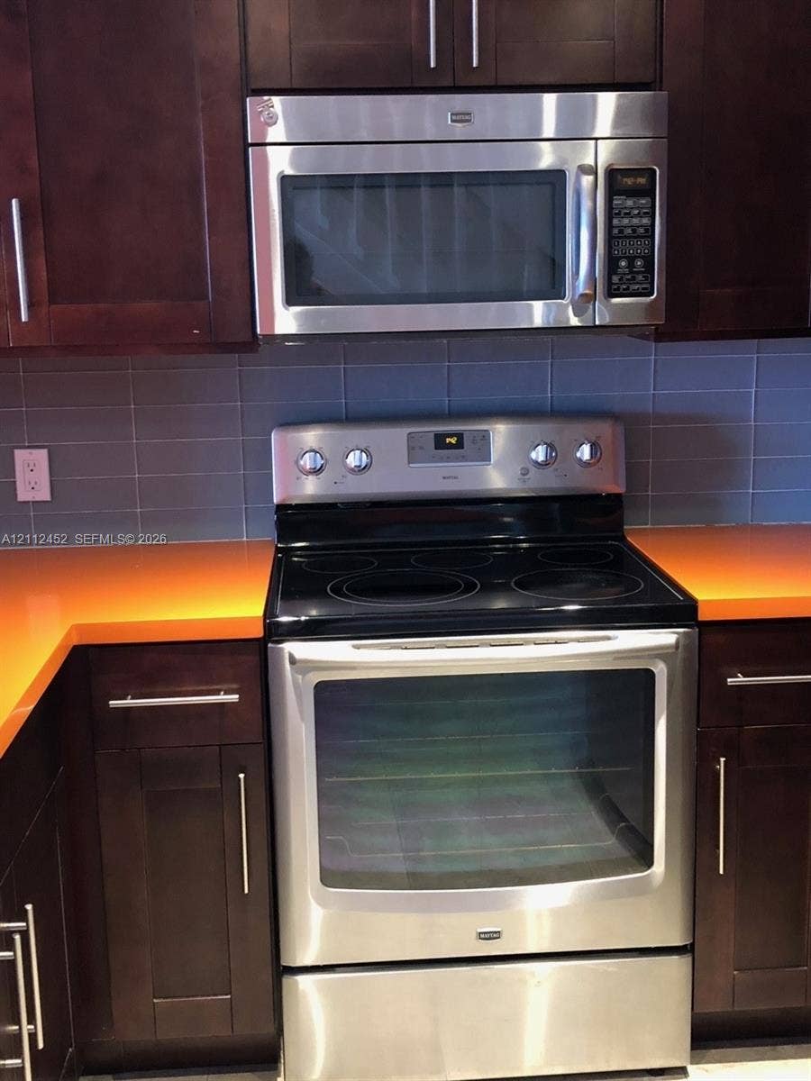 Dark-finish cabinetry with linear pulls, stainless steel over-the-range microwave, electric range with glass cooktop, solid-surface countertops, and horizontal subway tile backsplash