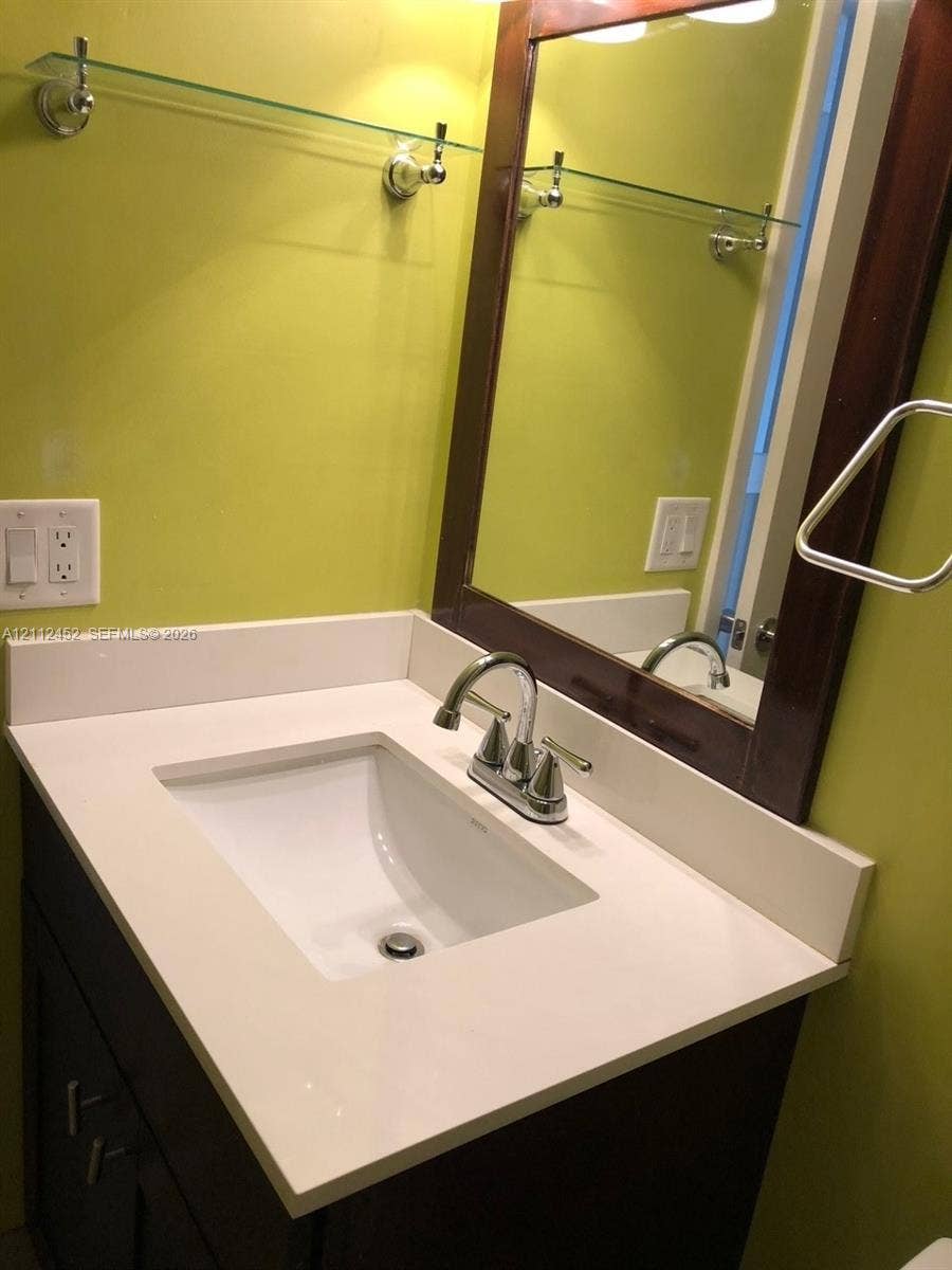 Dark wood-finish vanity with a white countertop and undermount sink, chrome faucet, framed mirror, and mounted glass shelving