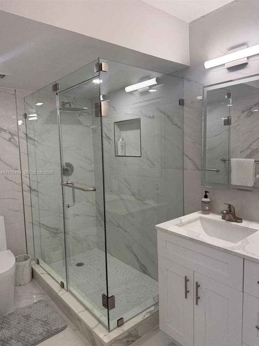 3800 S Ocean Drive, #1601, Hollywood, Hollywood, FL 33019 Walk-in shower featuring a frameless glass enclosure, recessed wall niche, and floor-to-ceiling veined stone-finish tile, complemented by a white vanity with a stone-finish countertop and an integrated mirror