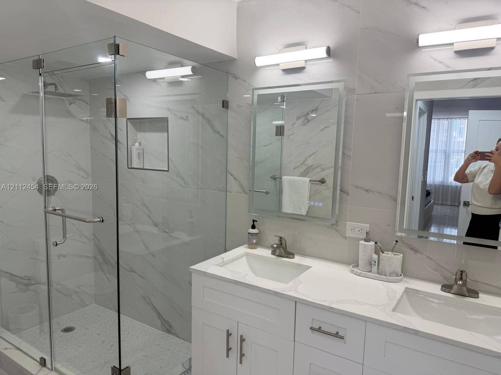 3800 S Ocean Drive, #1601, Hollywood, Hollywood, FL 33019 Dual-sink vanity with white cabinetry and stone countertop, glass-enclosed walk-in shower with integrated wall niche, floor-to-ceiling stone-style wall tile, and wall-mounted light fixtures