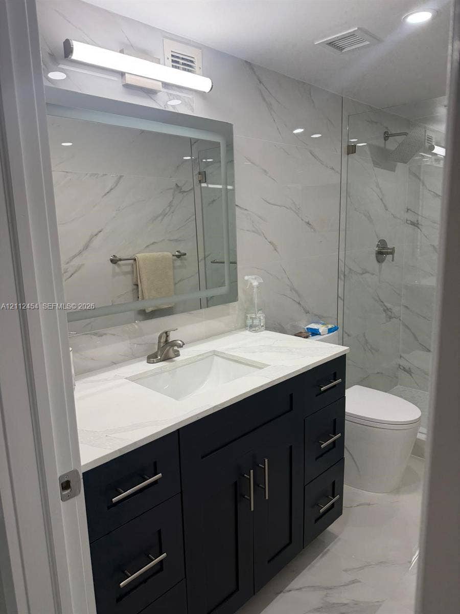 3800 S Ocean Drive, #1601, Hollywood, Hollywood, FL 33019 Dark-finish vanity with a rectangular sink, stone-surface countertop, LED-backlit mirror, and full-height stone-patterned wall tile