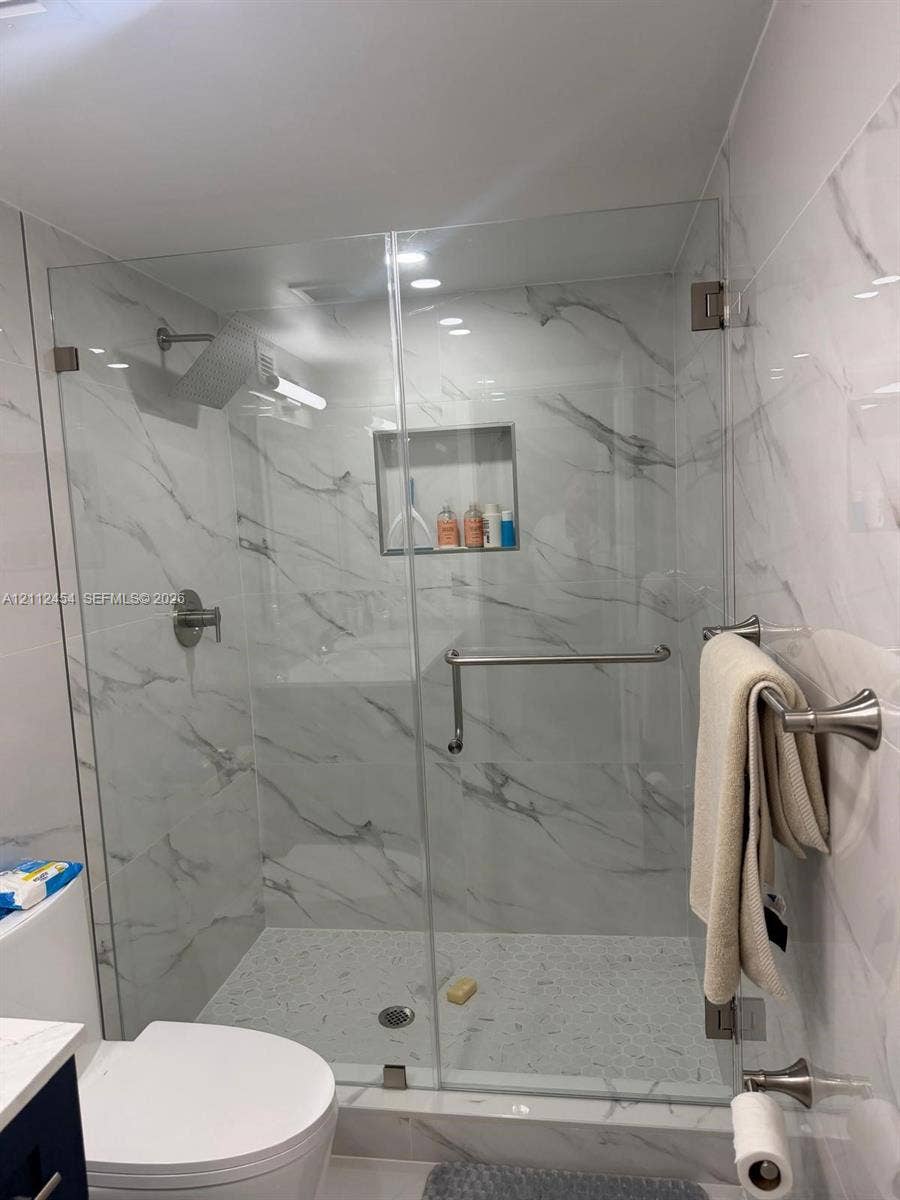 3800 S Ocean Drive, #1601, Hollywood, Hollywood, FL 33019 Walk-in shower featuring a frameless glass enclosure, floor-to-ceiling stone-pattern tile, integrated recessed shelving, polished metal hardware, and a pebble-style basin floor