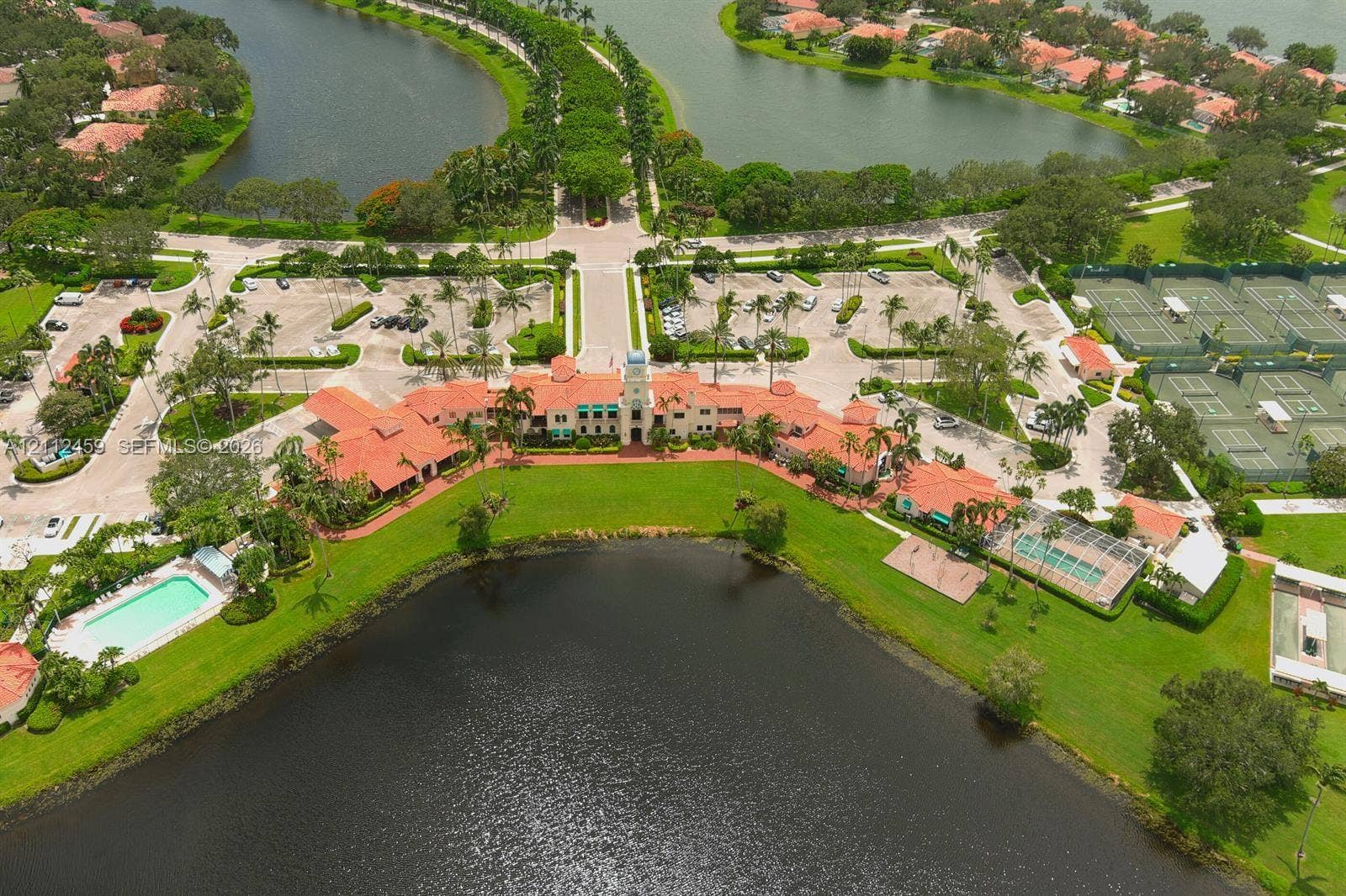 Aerial perspective of a clubhouse complex featuring a tile roof, waterfront positioning, multiple outdoor swimming pools, and tennis courts