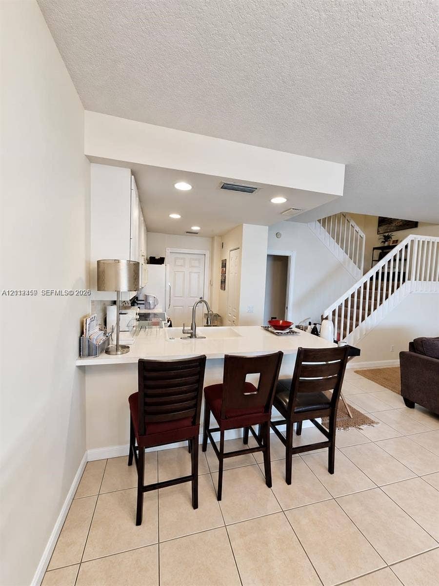 Kitchen featuring a breakfast bar with light-toned countertops, white cabinetry, a single-basin sink with a gooseneck faucet, and recessed lighting, accompanied by a staircase with white balusters