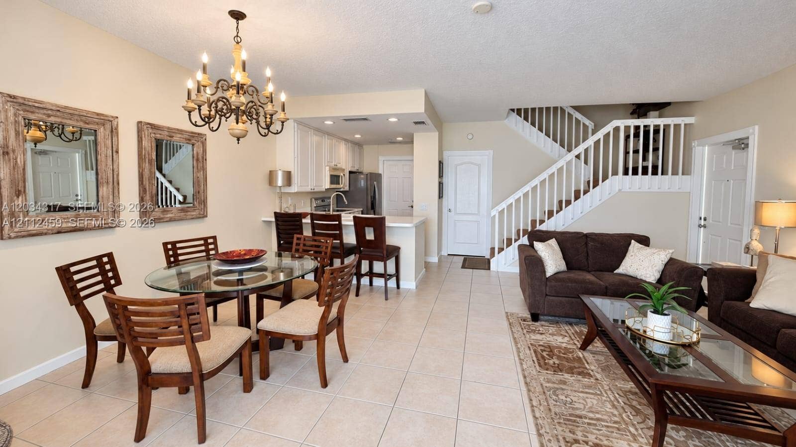 Open-concept area featuring tile flooring, kitchen breakfast bar with white cabinetry, multi-tiered metal chandelier, and white staircase with balustrades