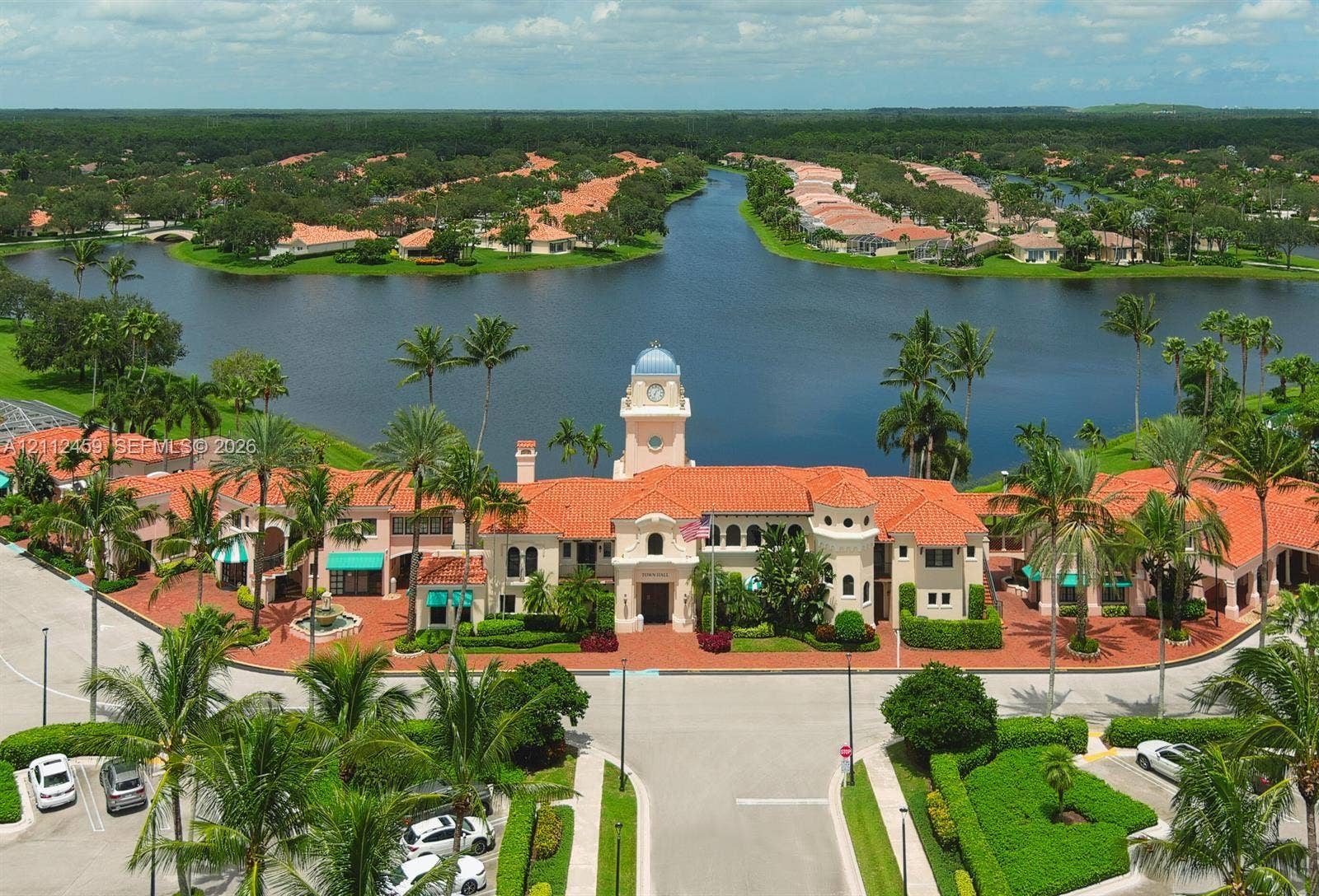 Mediterranean-style clubhouse featuring a central clock tower, terracotta tile roofing, stucco exterior, expansive water frontage, and brick-paved entryways