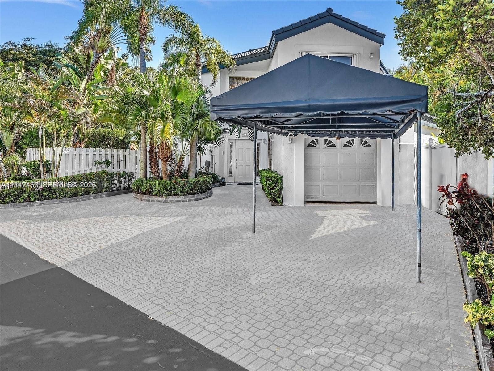 250 NE 212th Street, Miami, Miami, FL 33179 Exterior featuring a paved driveway, mature palm trees, multi-paneled garage door, and white privacy fencing