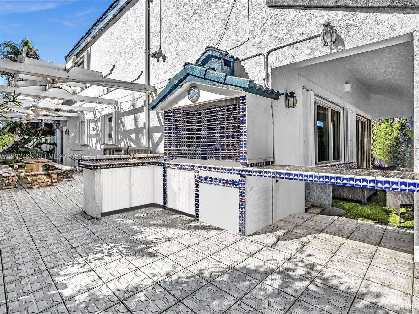 250 NE 212th Street, Miami, Miami, FL 33179 Outdoor kitchen featuring a built-in grill, tiled countertop, decorative patterned backsplash, and a gabled roof structure, complemented by a expansive tiled patio and a white wooden pergola