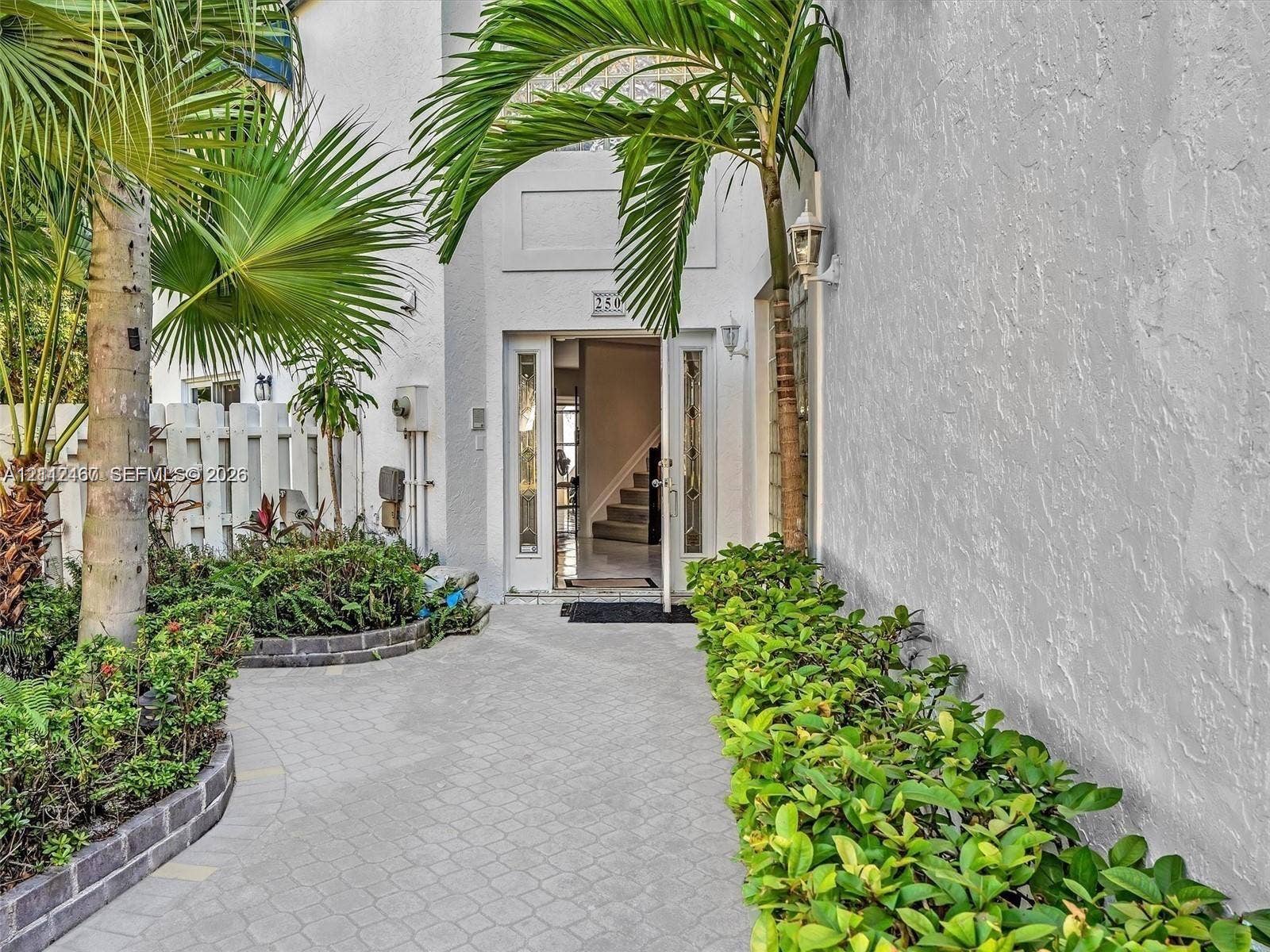 250 NE 212th Street, Miami, Miami, FL 33179 Stucco exterior entrance featuring a paver walkway, mature palm trees, decorative landscaping, and glass-panel door inserts