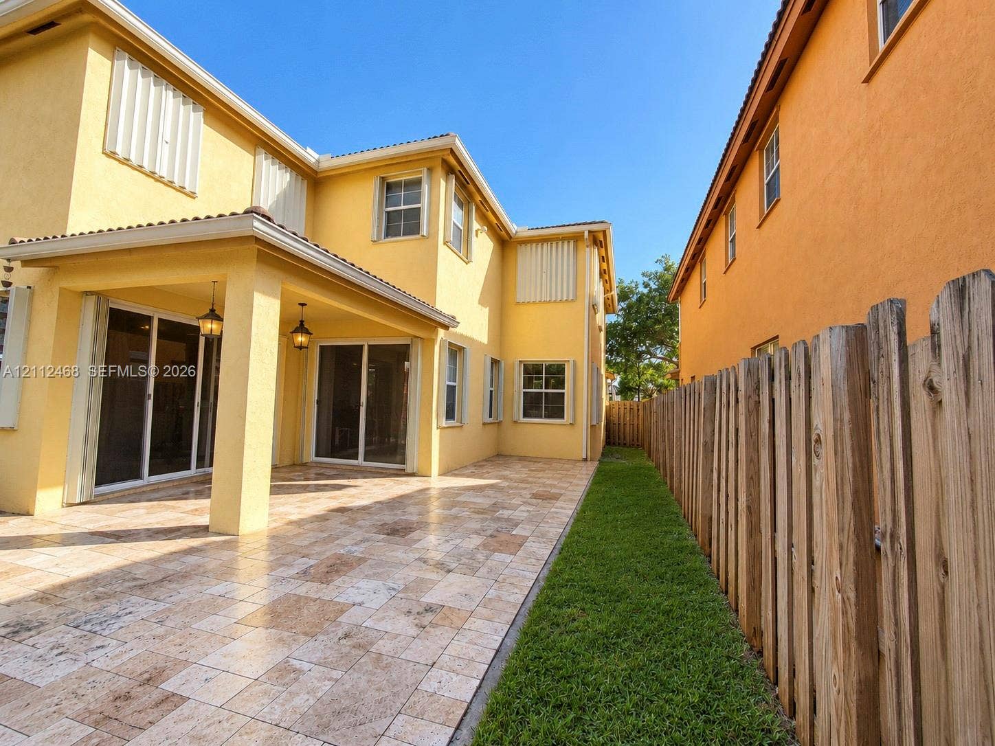 11009 NW 80th Lane, Doral, FL 33178 Stucco exterior featuring a covered patio with tile flooring, recessed outdoor lighting, dual sliding glass doors, and a wood-slat perimeter fence