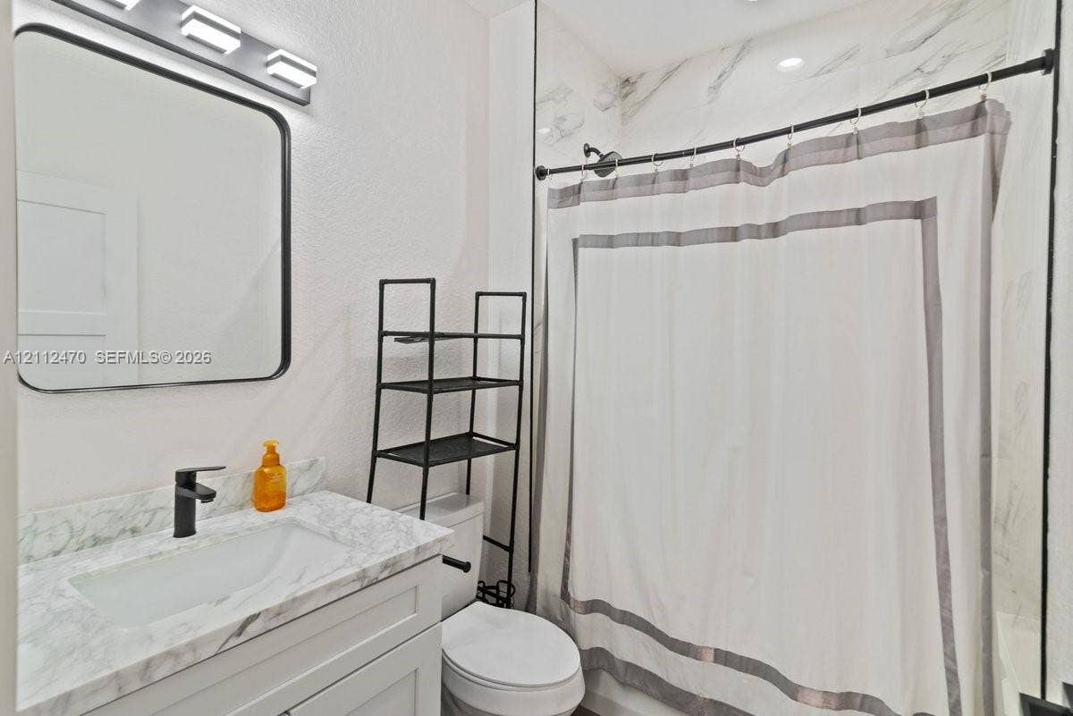 Bathroom vanity with stone-finish countertop, undermount sink, matte black faucet, rectangular mirror with a black frame, and shower surround with stone-finish wall tile