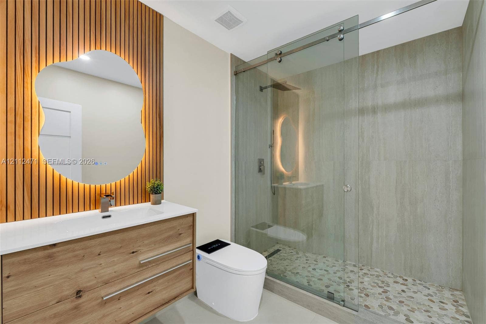 Wood-slat accent wall featuring a backlit, organic-shaped mirror, wood-finish vanity with an integrated basin, and a walk-in shower with a glass enclosure and pebble-stone flooring