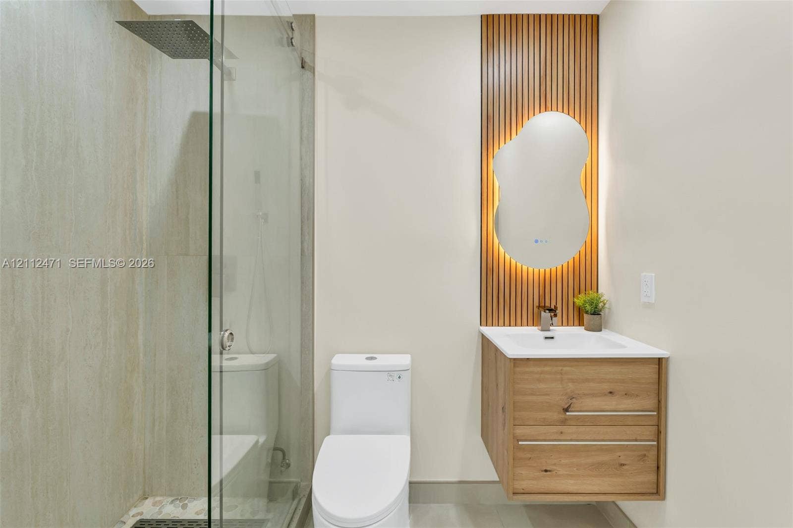 Walk-in glass enclosed shower with rainfall showerhead, floating wood-finish vanity, integrated solid surface sink, and decorative vertical wood-slat wall feature with backlit organic-shaped mirror