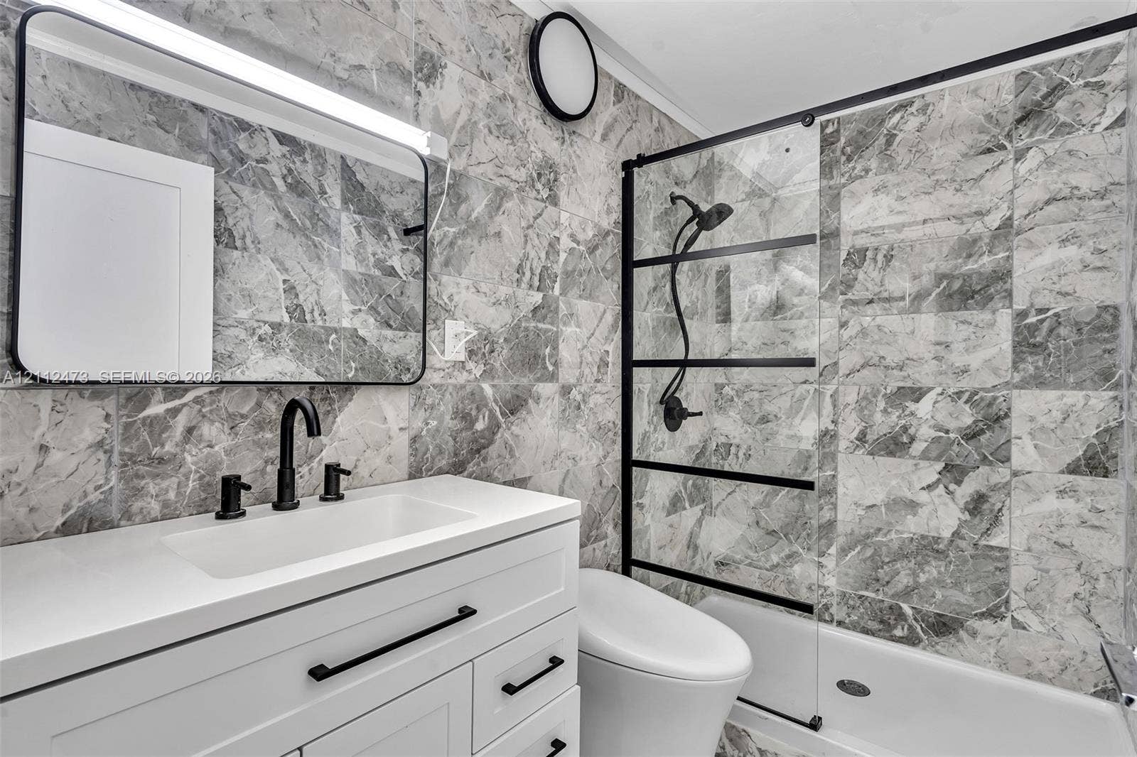 3340 SW 50th Street, Hollywood, Hollywood, FL 33312 Bathroom featuring full-height stone-finish tile surround, white vanity with quartz countertop, matte black fixtures, framed vanity mirror, and glass shower enclosure with black grid detailing
