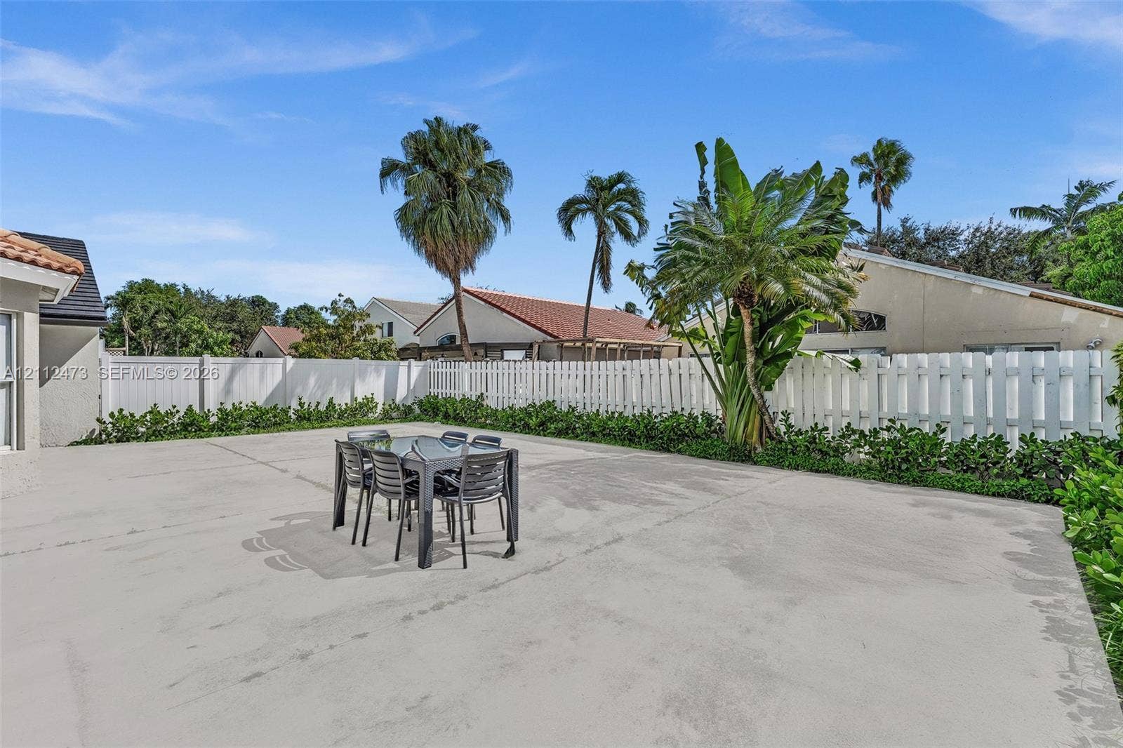 3340 SW 50th Street, Hollywood, Hollywood, FL 33312 Expansive concrete patio area enclosed by a white vinyl privacy fence, featuring mature palm trees and established perimeter landscaping