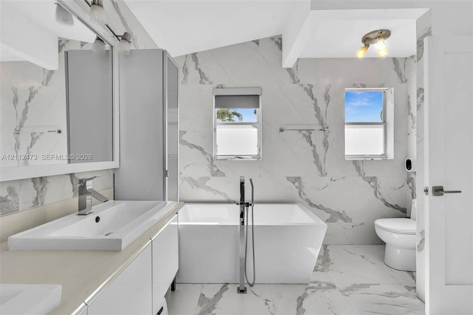 3340 SW 50th Street, Hollywood, Hollywood, FL 33312 Bathroom featuring floor-to-ceiling large-format tile with decorative veining, freestanding soaking tub with floor-mounted faucet, rectangular vessel sink atop a modern vanity, and dual wall-mounted light fixtures