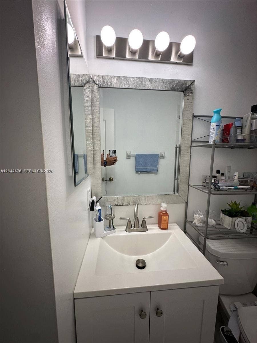 White vanity featuring a square basin, brushed metal centerset faucet, and framed vanity mirror topped with a four-bulb brushed metal light fixture