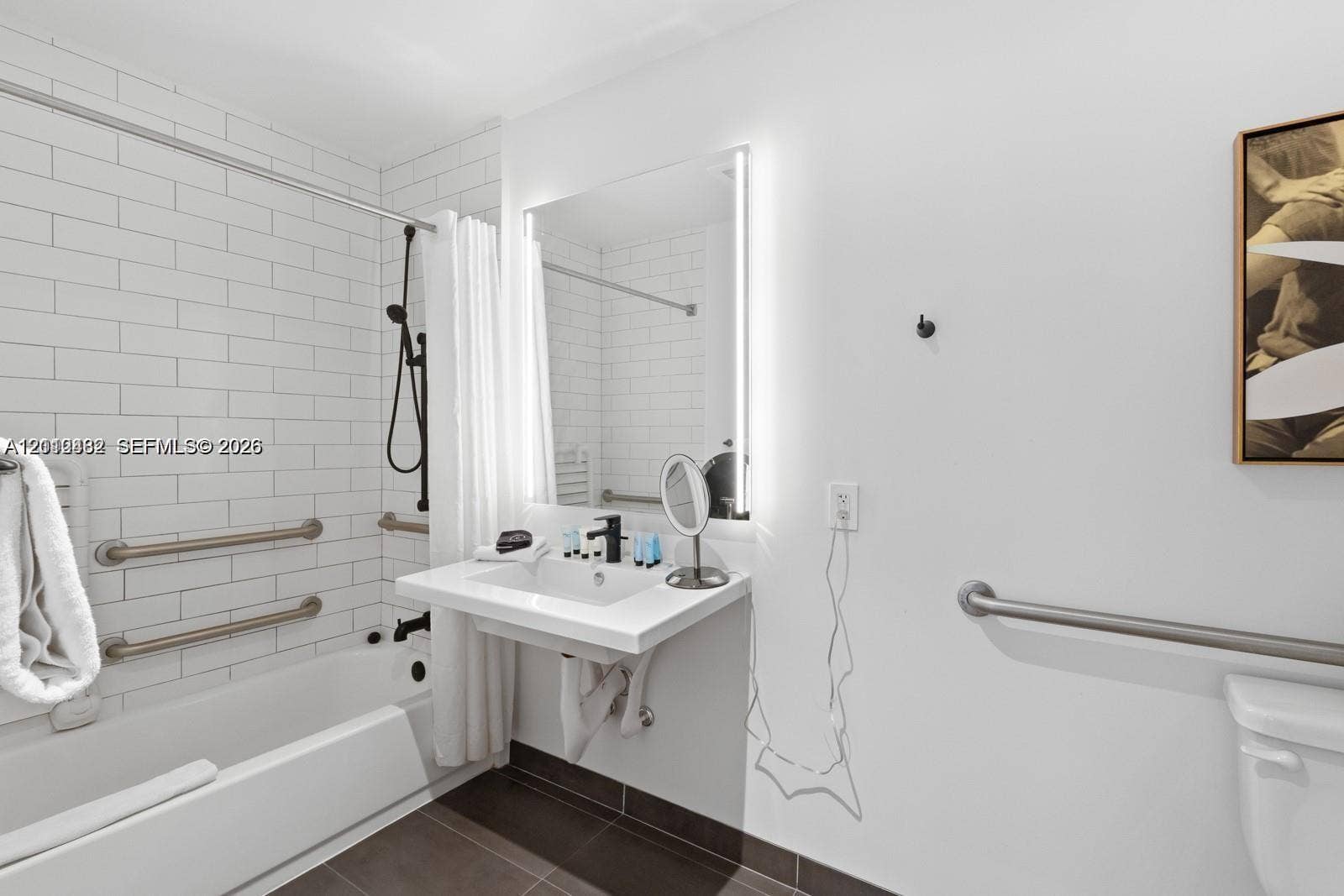 Wall-mounted floating vanity with integrated sink, rectangular illuminated mirror, white subway-tiled shower surround, matte black plumbing fixtures, and installed safety grab bars
