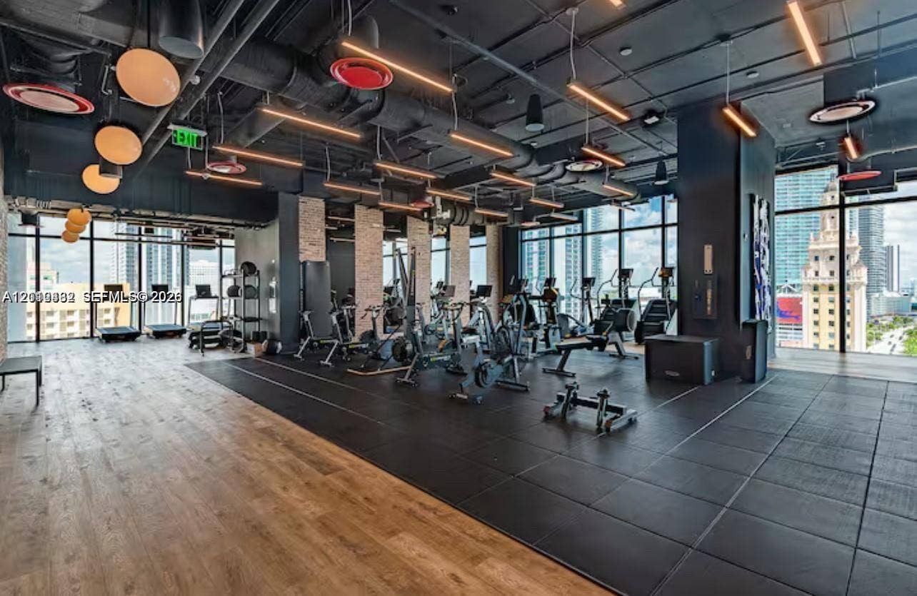 Industrial-style fitness center featuring floor-to-ceiling windows, exposed structural ceiling, wood-finish flooring, rubberized workout surfacing, and decorative globe lighting