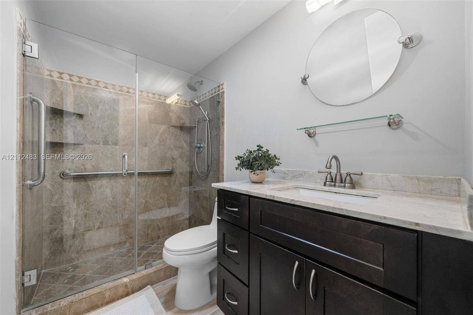 3505 S Ocean Drive, #1101, Hollywood, FL 33019 Dark wood vanity with a stone countertop, frameless glass shower enclosure, stone-tiled shower surround with built-in shelving, adjustable handheld shower head, and an oval wall-mounted mirror