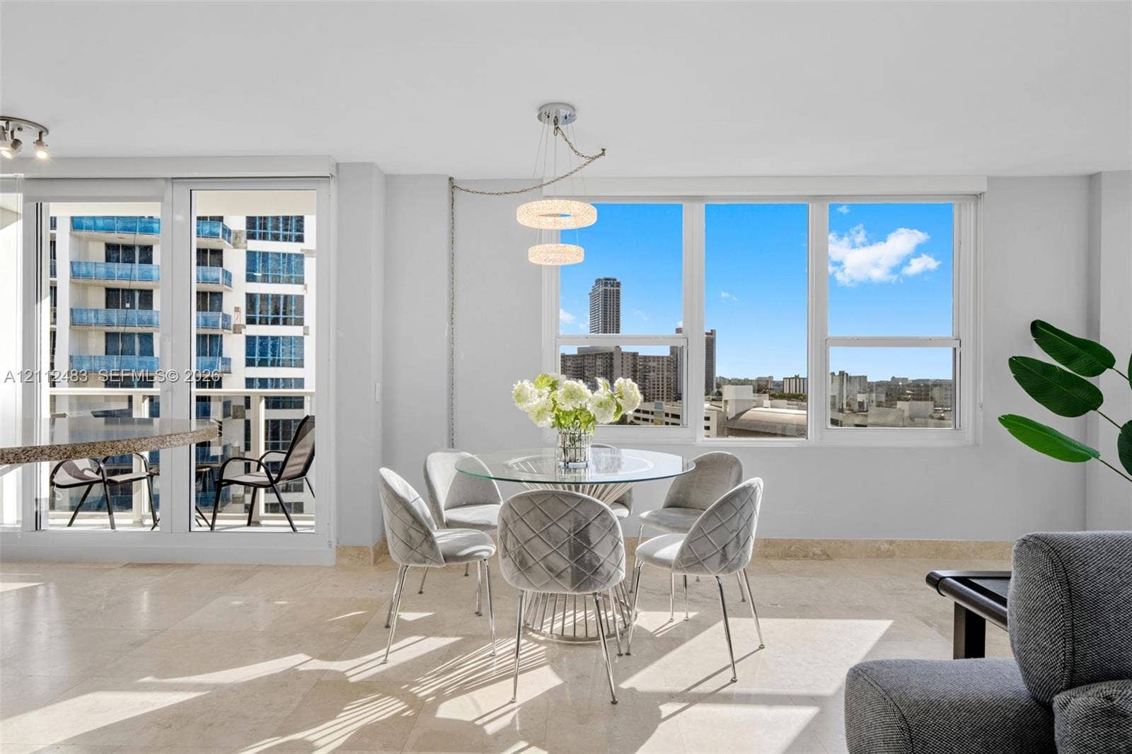 3505 S Ocean Drive, #1101, Hollywood, FL 33019 Light-filled interior featuring floor-to-ceiling windows, natural stone flooring, a multi-tier contemporary chandelier, and a glass-paneled door opening to a private balcony
