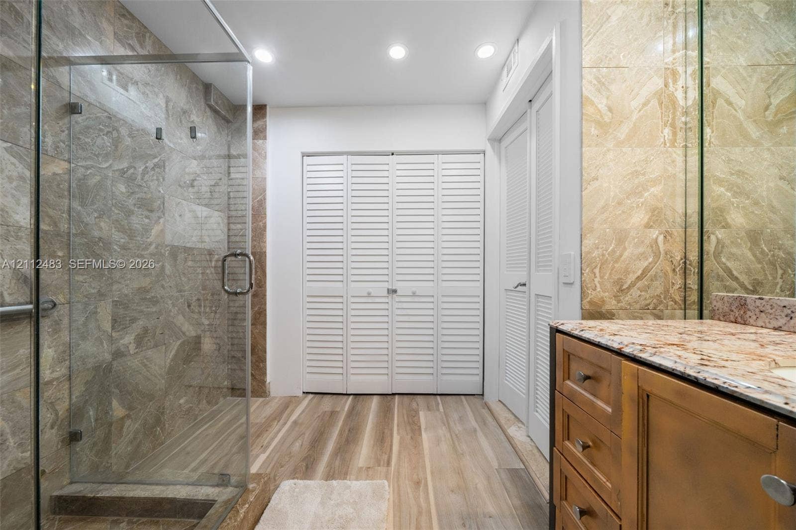 3505 S Ocean Drive, #1101, Hollywood, FL 33019 Walk-in shower with stone tile surround and glass enclosure, wood-finish flooring, stone-topped vanity with built-in cabinetry, and white louvered closet doors