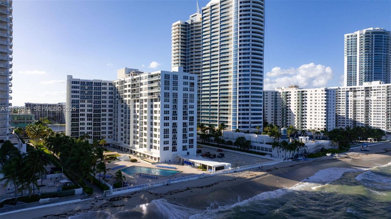 3505 S Ocean Drive, #1101, Hollywood, FL 33019 High-rise residential building featuring expansive beachfront frontage, an outdoor swimming pool, paved surface parking, and mature palm trees