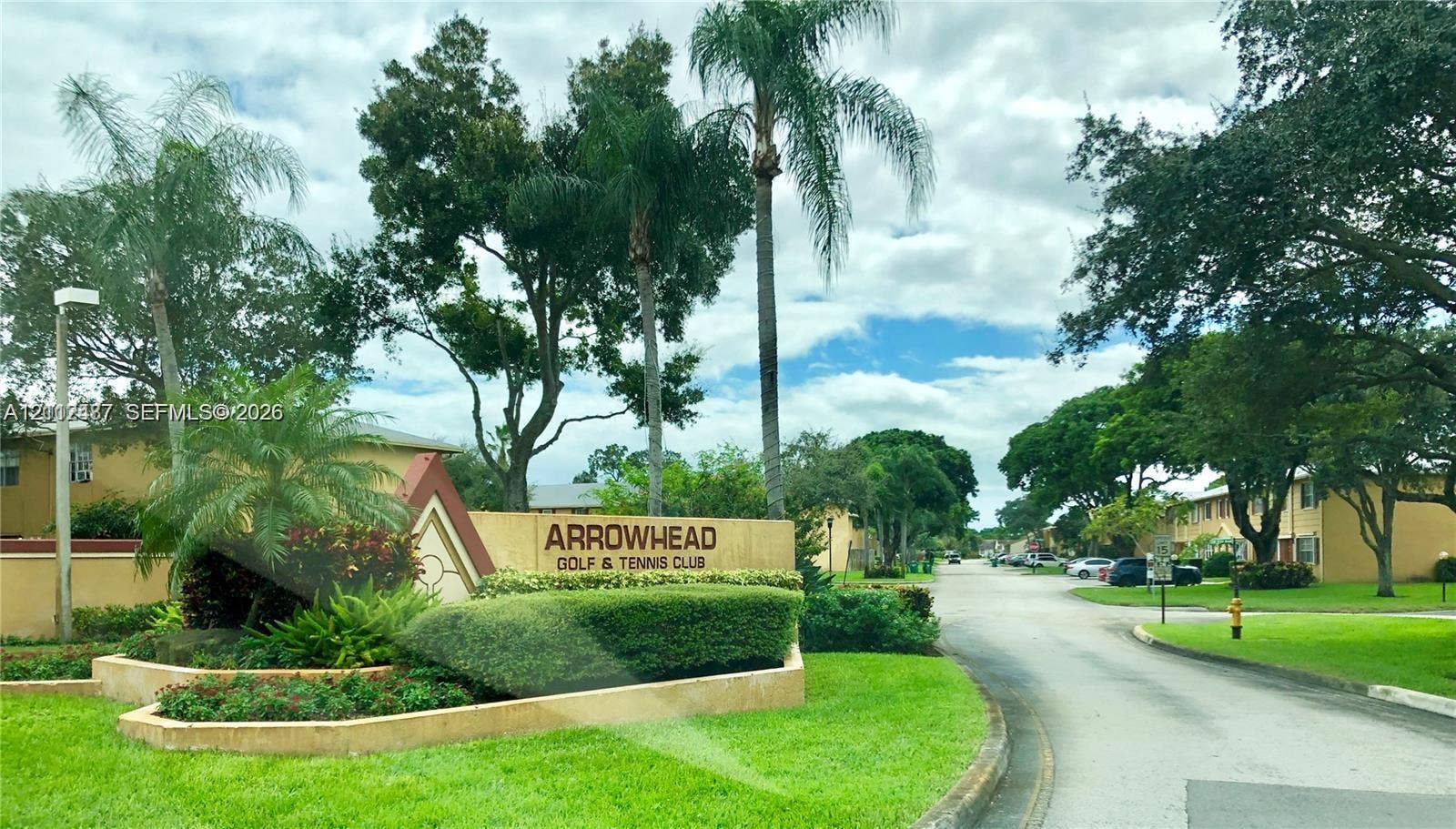 Community entrance featuring stucco monument signage, manicured landscaping with tropical palm trees, paved roadway, and mature canopy trees