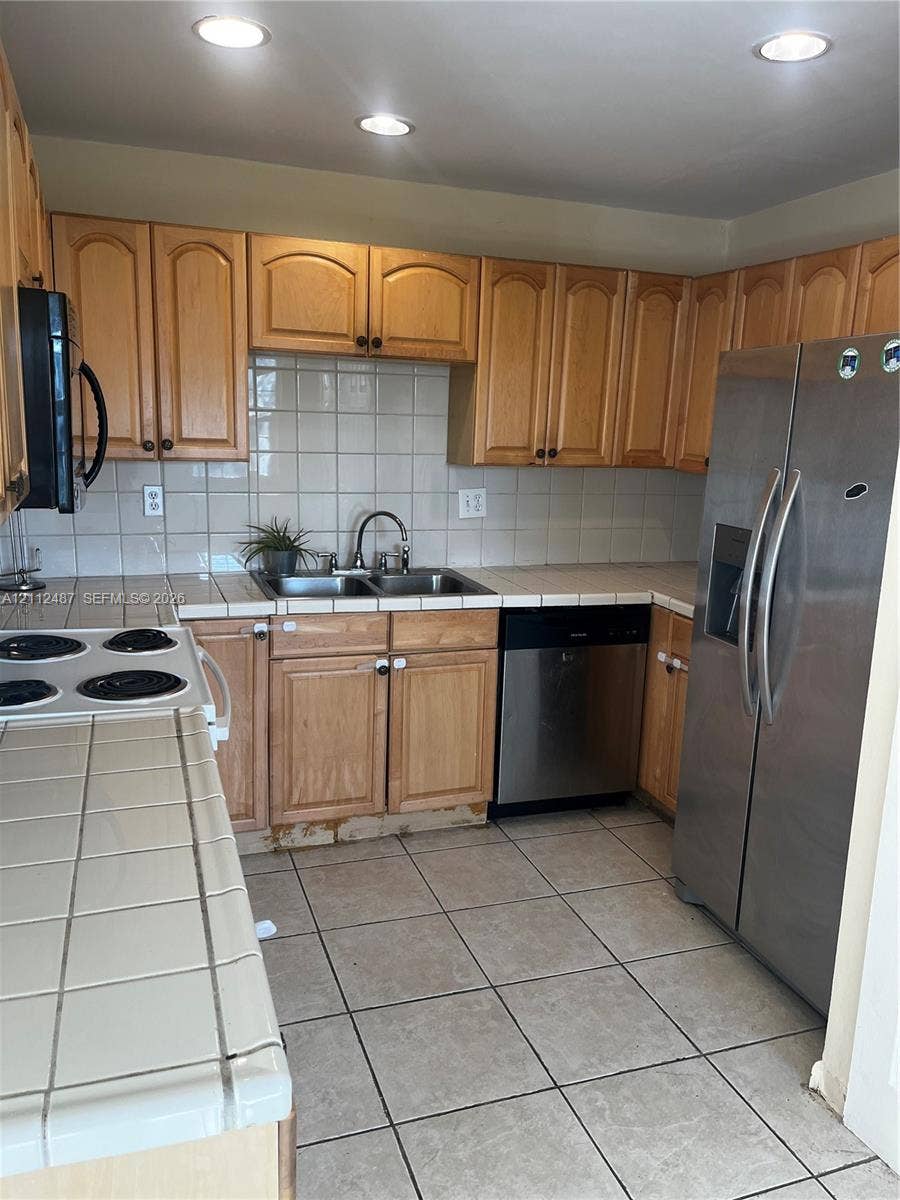 Kitchen featuring light wood cabinetry, tiled countertops and backsplash, stainless steel refrigerator and dishwasher, and ceramic tile flooring