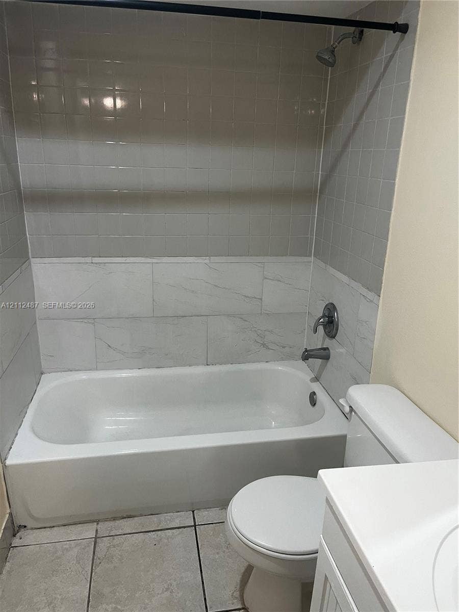 Full bathroom featuring a white porcelain tub with a two-tone tiled surround, integrated shower fixture, tile flooring, and a white vanity