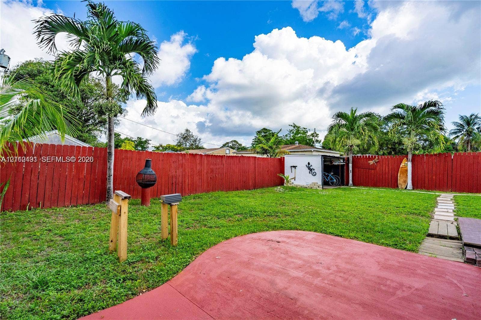 2509 Grant Street, Hollywood, FL 33020 Backyard featuring a red-painted concrete patio, mature palm trees, a grass lawn, a wood-slat privacy fence, and a detached storage shed