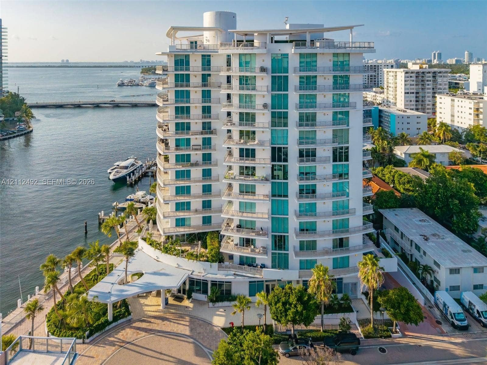 High-rise waterfront building featuring floor-to-ceiling glass windows, private balconies, a porte-cochre entryway, and tropical landscaping
