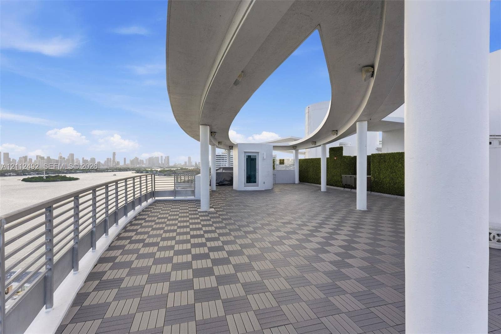 Expansive outdoor terrace featuring a geometric tile deck, curved architectural overhang, cylindrical support columns, metal railing, and privacy hedging
