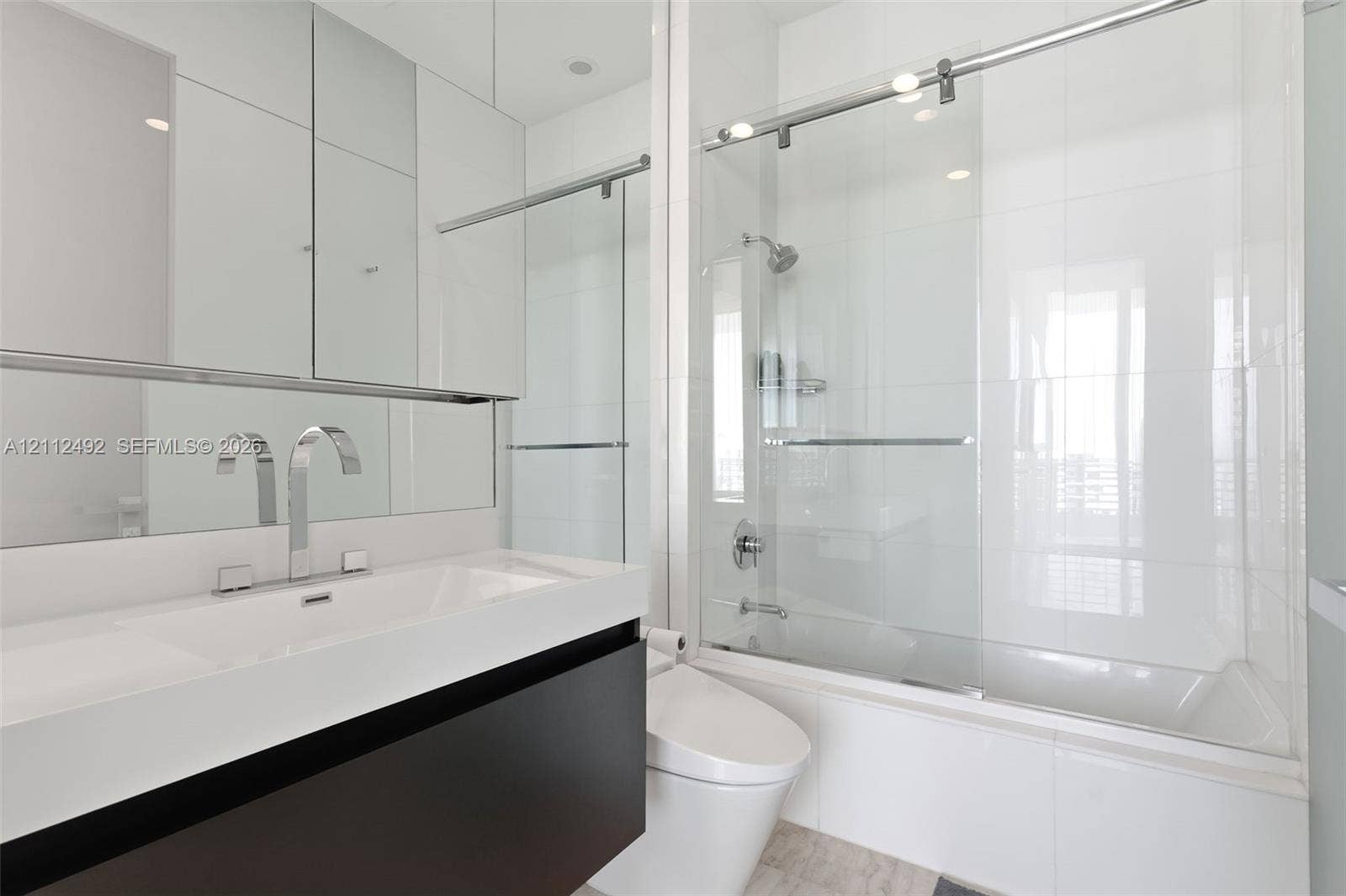 Modern vanity with integrated rectangular basin and polished chrome widespread faucet, mirrored medicine cabinet, floor-to-ceiling tiled shower enclosure with sliding glass door, and wall-mounted commode