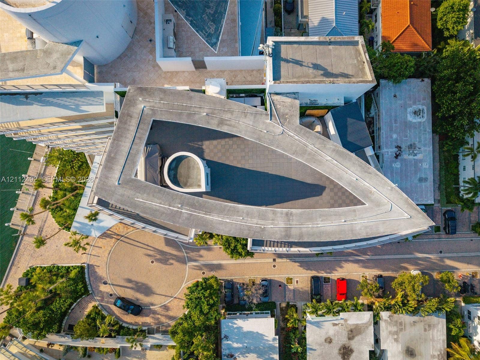Aerial perspective of a multi-level structure featuring a distinctive triangular roofline, a circular driveway, integrated hardscaping, and immediate proximity to a waterway with boat dockage