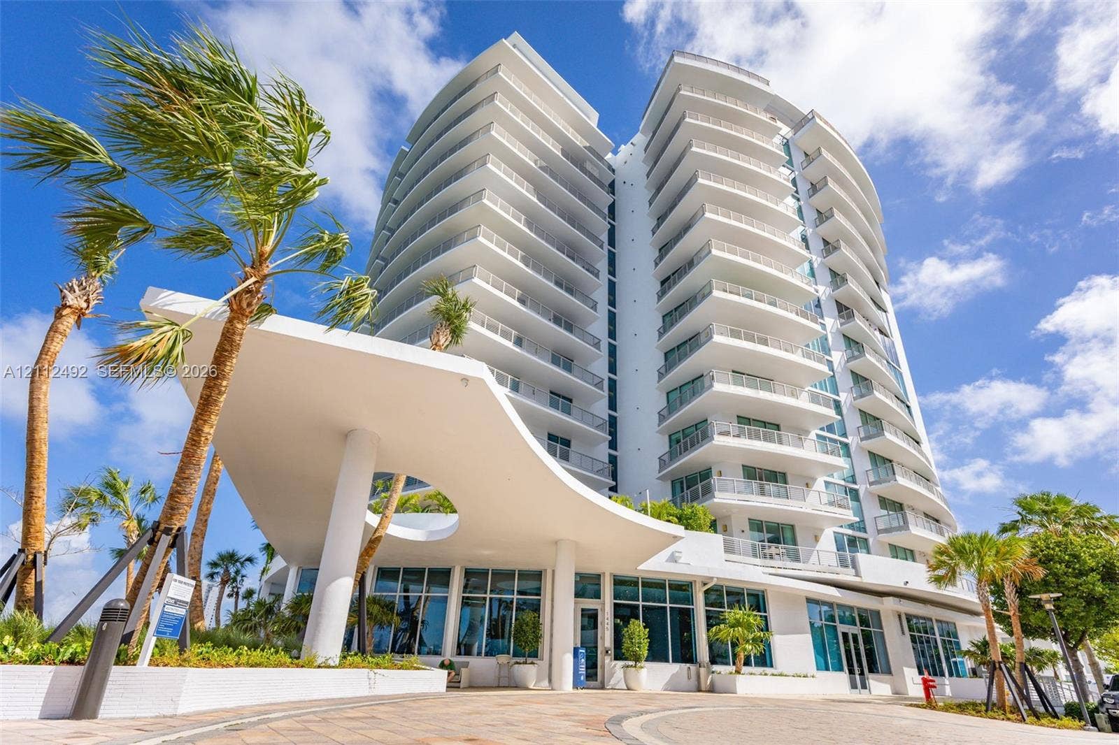 High-rise residential building featuring a sculpted architectural porte-cochre, stacked private balconies, floor-to-ceiling windows, and paved motor court