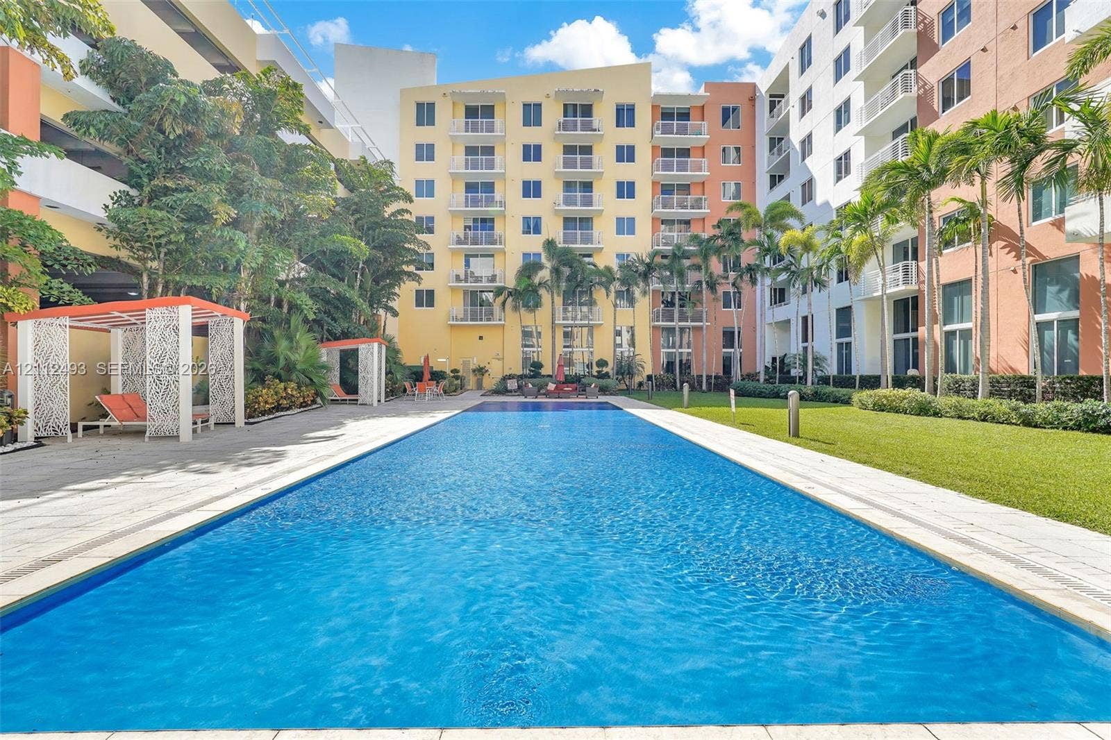 Rectangular swimming pool surrounded by stone paver decking, manicured lawn, architectural cabanas, and mature palm trees