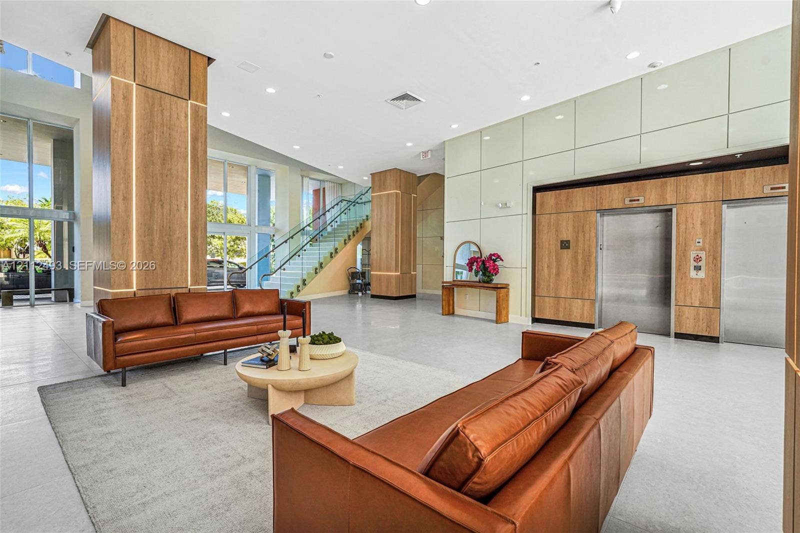 Grand lobby featuring floor-to-ceiling wood-finish pillar detailing, polished tile flooring, a glass-panel staircase, and dual stainless steel elevators