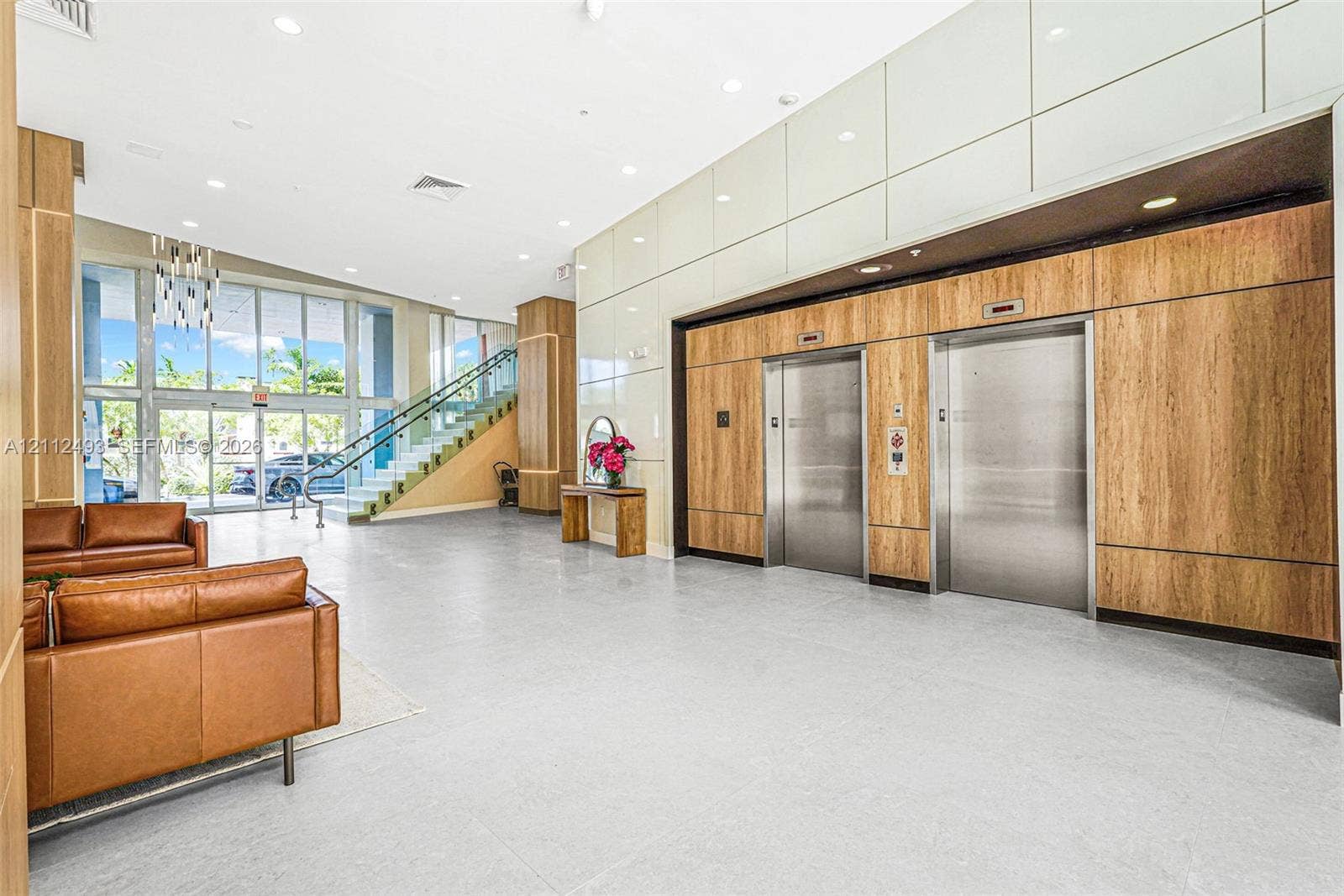 Expansive lobby featuring light-toned tile flooring, floor-to-ceiling wood-finish wall paneling, dual stainless steel elevators, and floor-to-ceiling glass windows with a staircase
