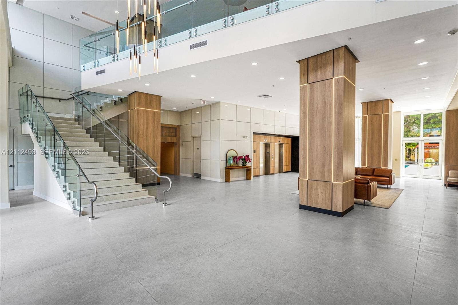 Grand lobby featuring expansive tile flooring, a glass-railed staircase, integrated wood-paneled pillars with accent lighting, and a multi-tier sculptural chandelier