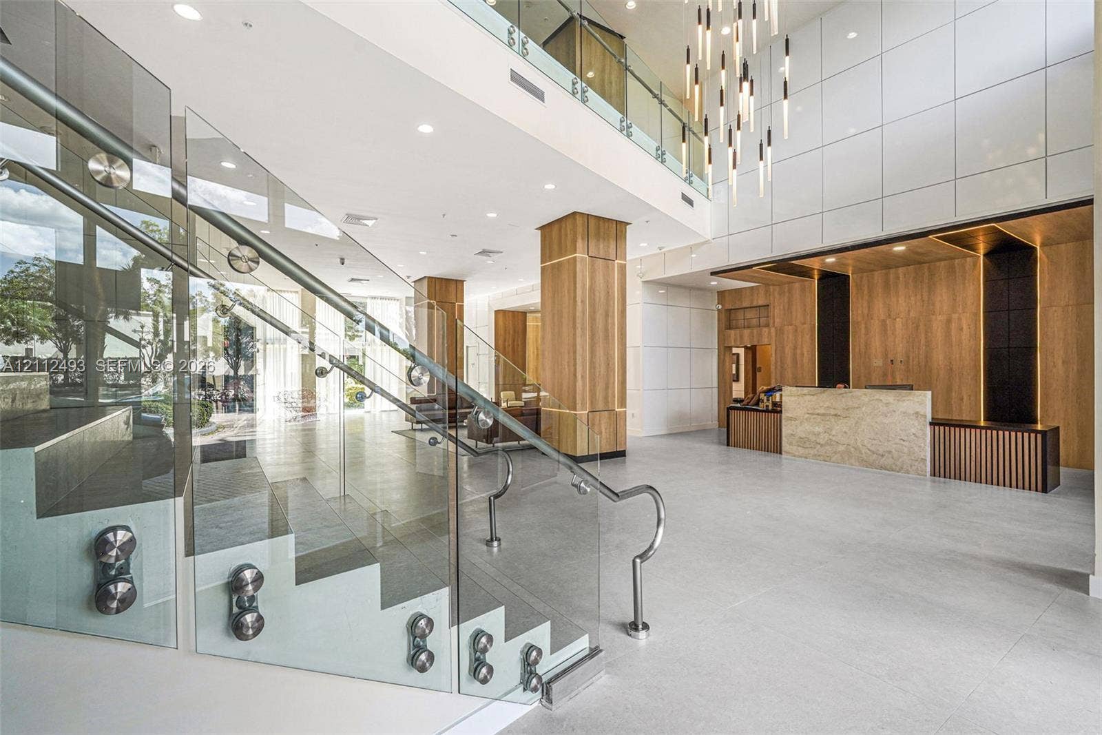 Grand atrium featuring a multi-story open staircase with glass railings and metal hardware, floor-to-ceiling paneling, large-format tile flooring, a stone-front reception desk, and a tiered modern chandelier