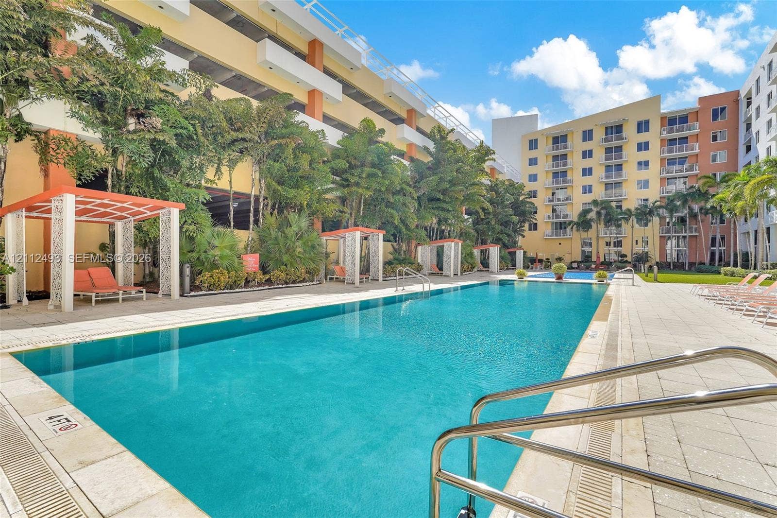 Swimming pool featuring a tiled deck, metal handrails, integrated shade structures, and surrounding palm trees