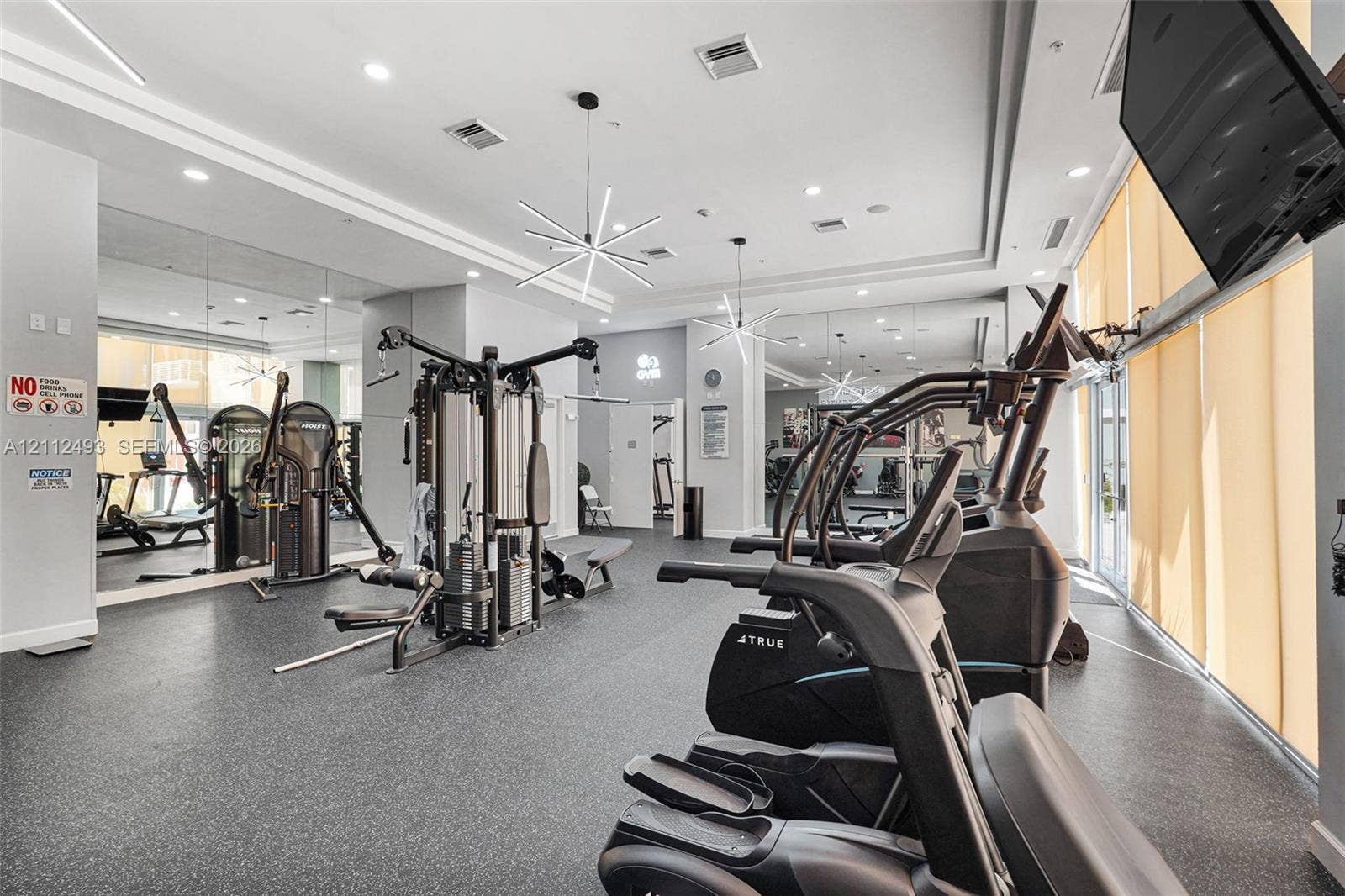 Fitness center featuring multi-station exercise equipment, recessed ceiling lighting, geometric pendant light fixtures, wall-to-wall mirrors, and floor-to-ceiling windows with roller shades