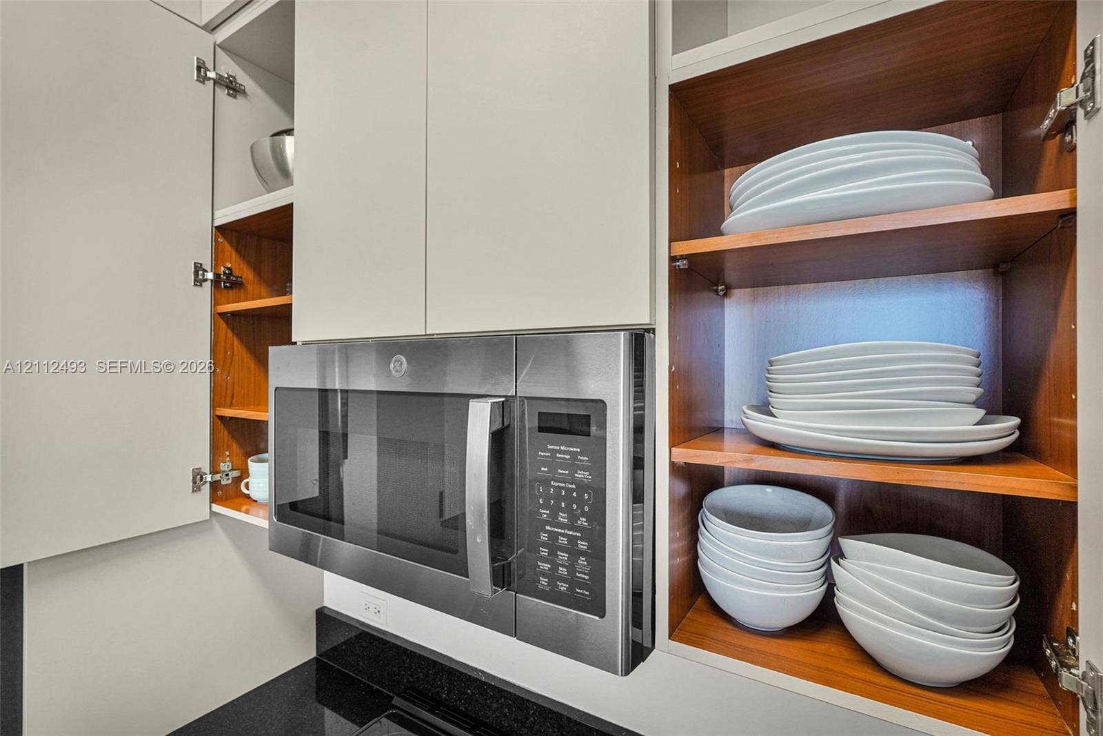 Built-in stainless steel microwave, matte white upper cabinetry, exposed wood-finish shelving, and black solid-surface countertop
