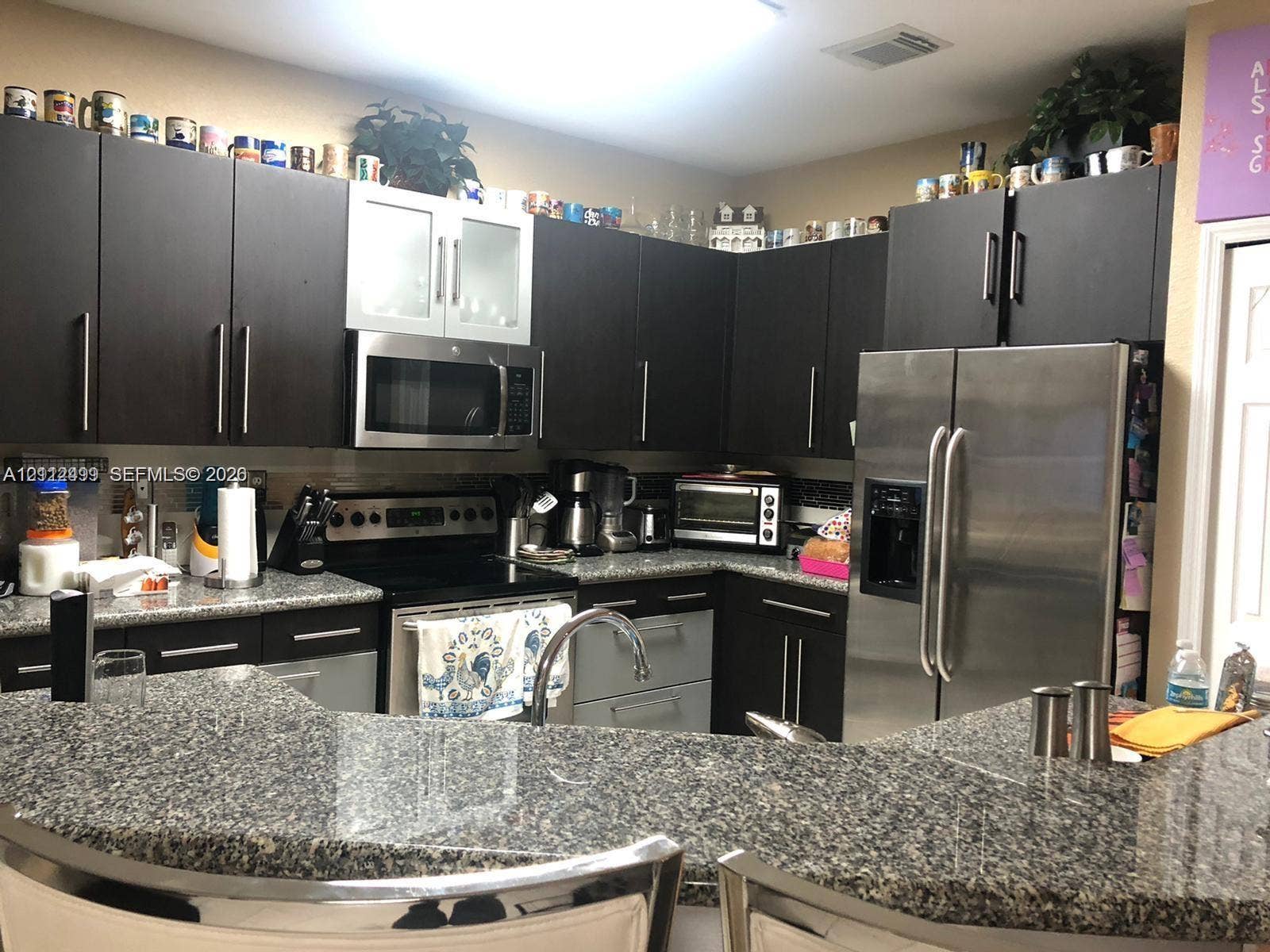 Kitchen featuring dark cabinetry, stone countertops, stainless steel appliances, a microwave, and a peninsula with a built-in sink