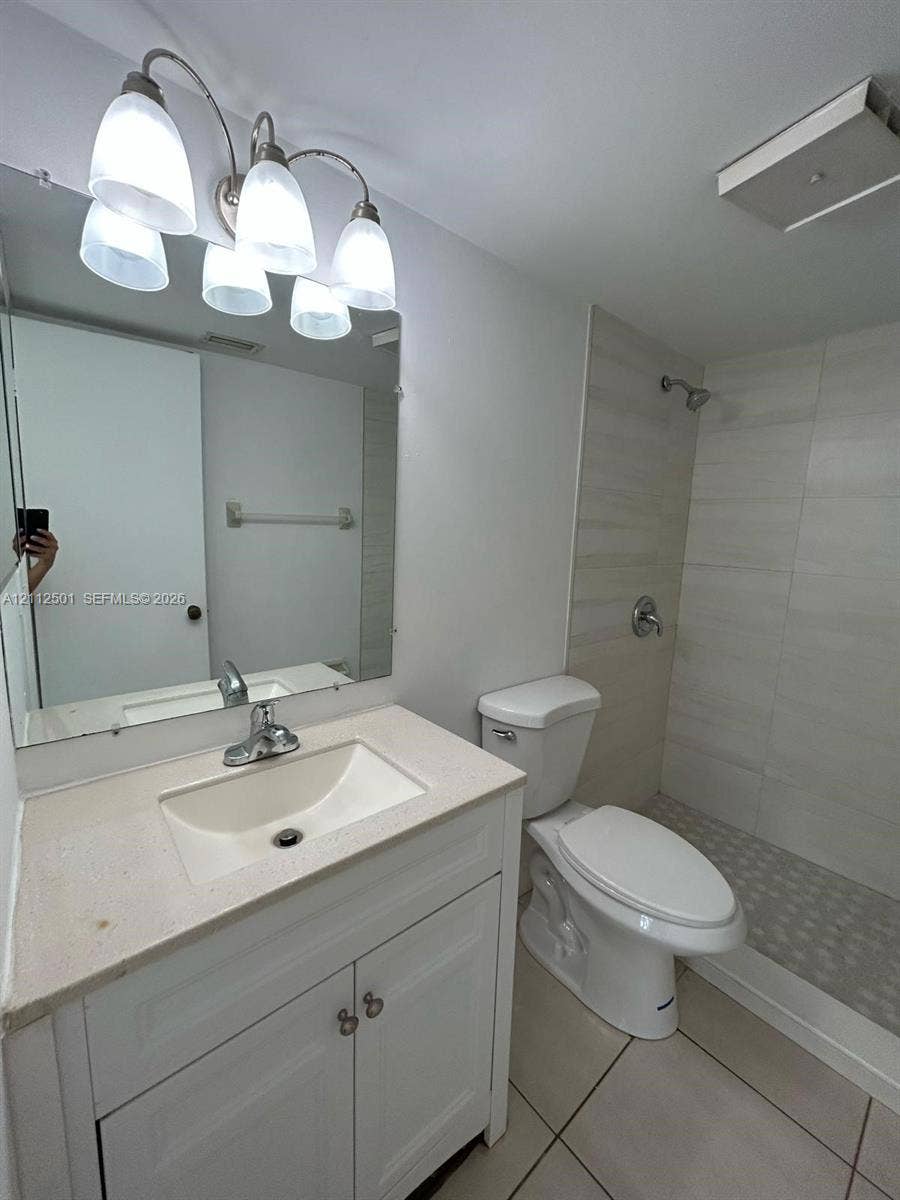 Bathroom featuring a white vanity cabinet with a light-toned countertop, a multi-light fixture, a wall-mounted mirror, a walk-in shower with tile surround, and tile flooring