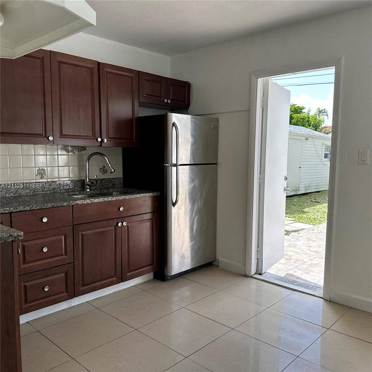 Kitchen featuring dark wood cabinetry, stone-finish countertops, ceramic tile backsplash, stainless steel refrigerator, and large-format tile flooring
